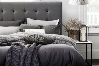 Bed with gray tufted headboard, layered pillows, cozy blankets, decorative pillow, and nightstand with branches and candle.