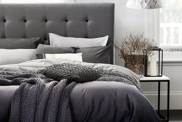 Bed with gray tufted headboard, layered pillows, cozy blankets, decorative pillow, and nightstand with branches and candle.