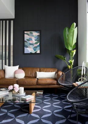 Stylish living room with brown leather sofa, abstract art, glass coffee table, black chairs, and large houseplant.