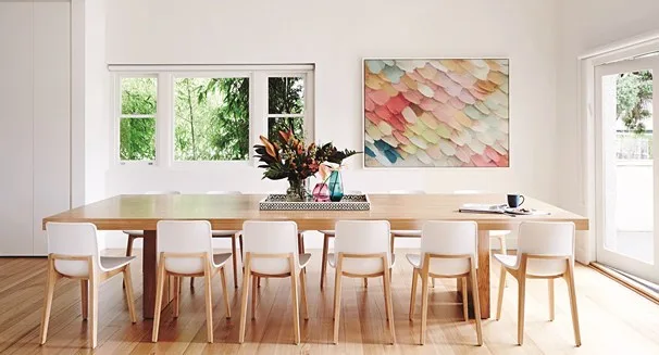 Spacious dining room with a long wooden table, white chairs, floral centerpiece, and abstract wall art under natural light.