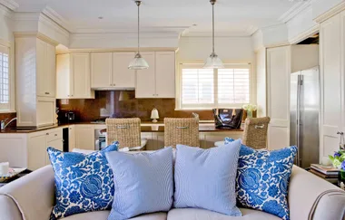 A modern kitchen with cream cabinets, two pendant lights, wicker chairs, and a couch with blue patterned pillows.