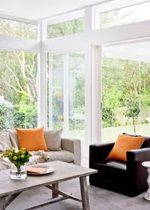 Bright living room with large windows, two armchairs with orange pillows, wooden table, and greenery outside.