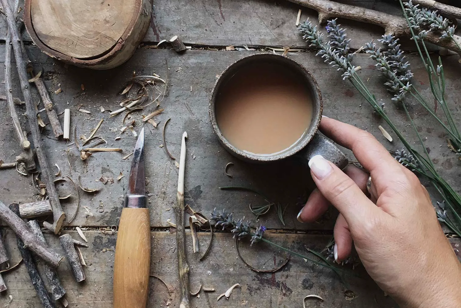 Hand holding a mug of coffee on a wooden table with carving tools, wood shavings, and lavender sprigs.