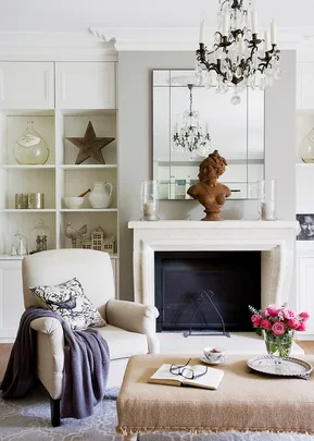 Living room with armchair, cozy blanket, and decorative items; a chandelier hangs above a mantel with a bust sculpture.