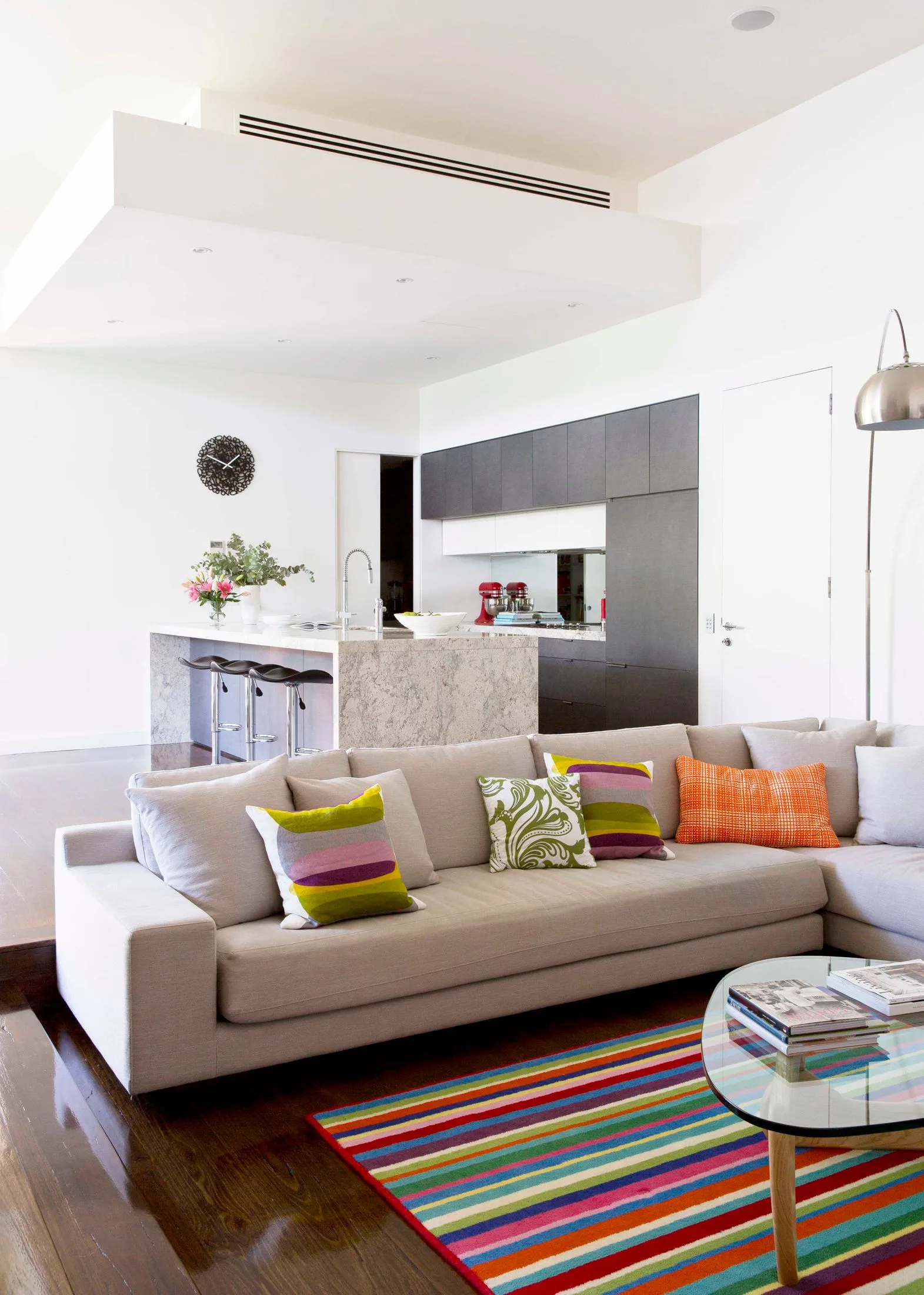 Contemporary living room with a beige sofa, colorful cushions, striped rug, and a modern kitchen in the background.