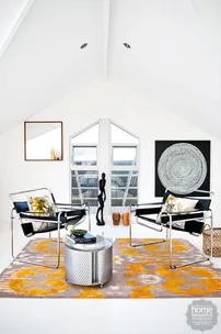 Modern living room with two chairs, round table, abstract art, and bright yellow patterned rug under high ceiling.
