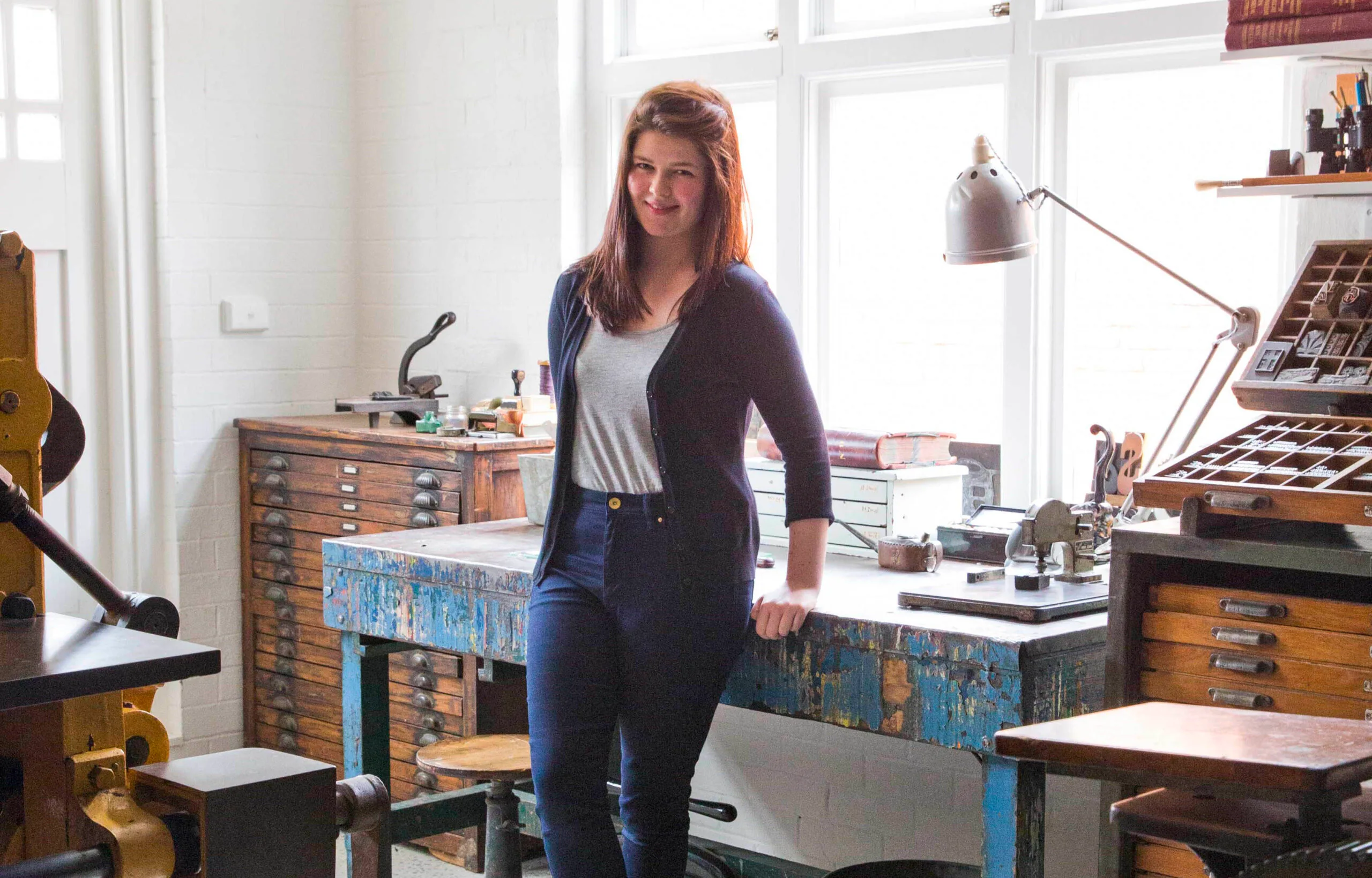 A woman stands by a wooden desk in a well-lit workshop with art supplies and books.