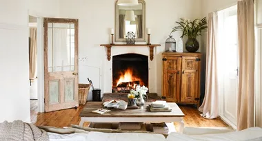 A cozy living room with a lit fireplace, wooden furniture, and rustic decor, featuring a large mirror above the mantel.