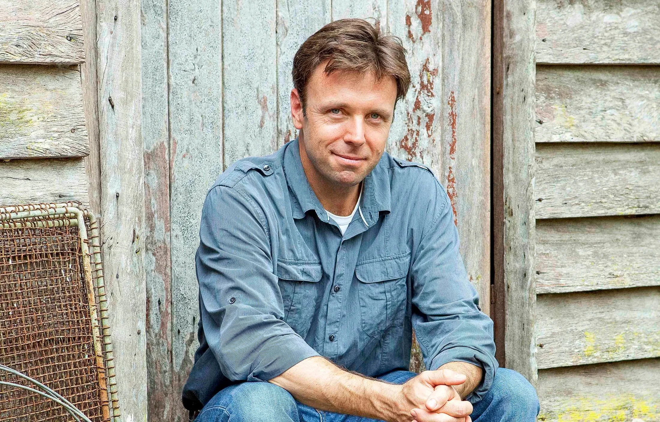 Man in a blue shirt sitting in front of a rustic, weathered wooden background.