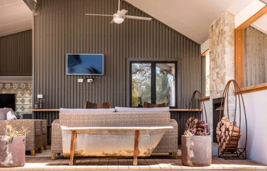 Outdoor patio with wicker furniture, a wall-mounted TV, firewood, and potted plants on a wooden deck.