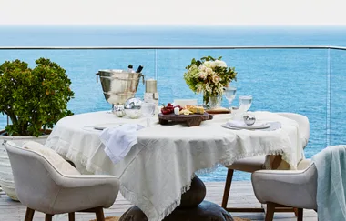 Elegant outdoor table setup with white cloth, flowers, a cheese platter, and champagne overlooking the ocean.