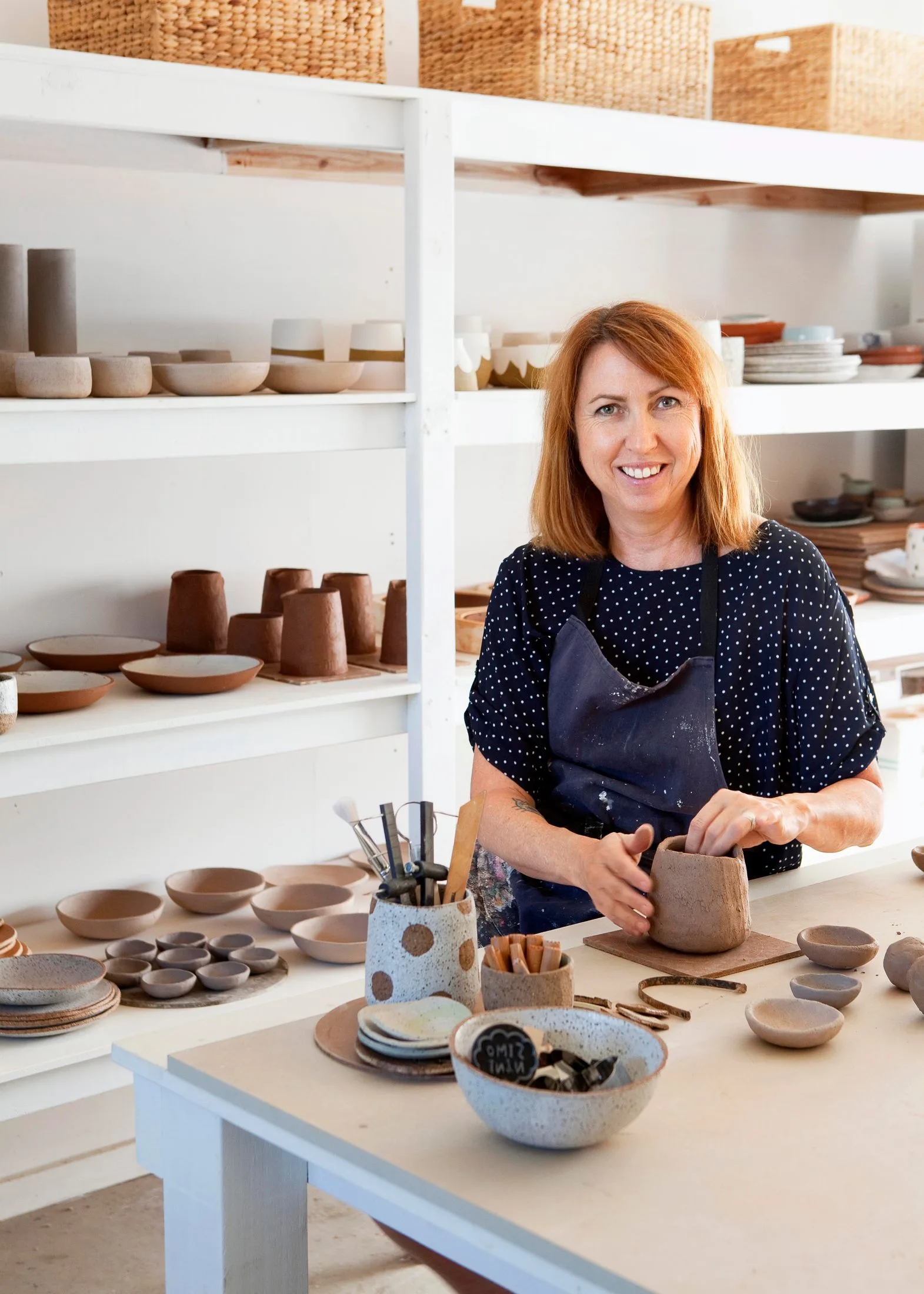 Potter in a studio shaping clay, surrounded by bowls and pottery on shelves, with brushes and tools on the table.