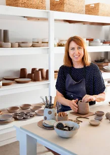 Potter in a studio shaping clay, surrounded by bowls and pottery on shelves, with brushes and tools on the table.
