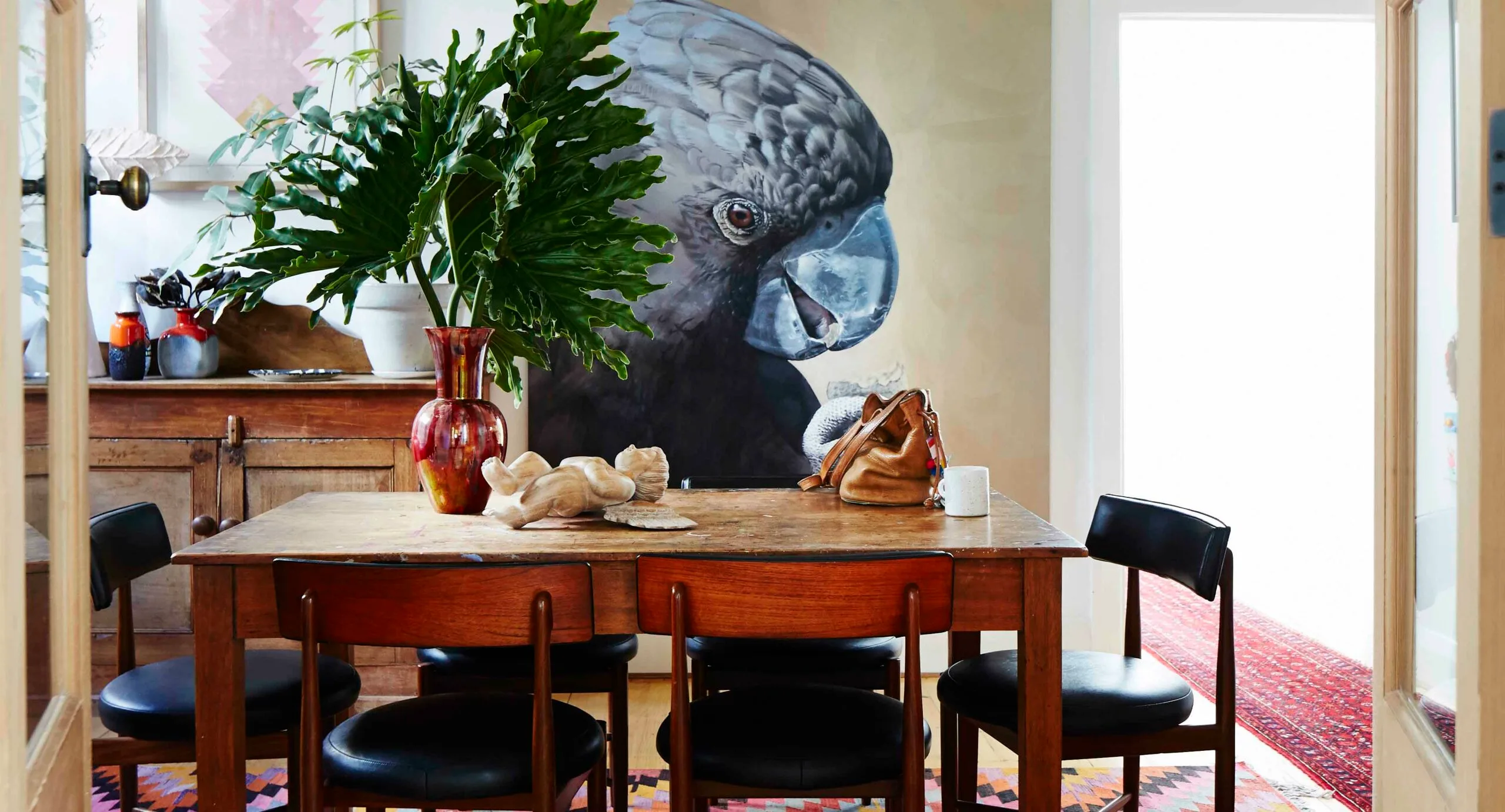 Dining room with a wooden table, black chairs, large plant, and a mural of a black cockatoo on the wall.