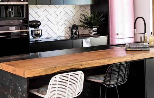 Modern kitchen with black cabinets, herringbone backsplash, wooden island, pink fridge, and two bar stools.