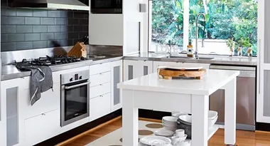 Modern white kitchen with stainless steel appliances, island, window view of greenery, and wooden floor.