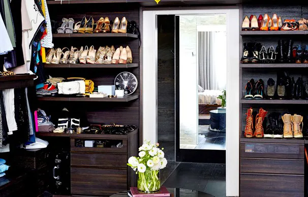Luxurious walk-in closet with shelves of shoes, handbags, a clock, and a vase of flowers. Doorway leads to a bedroom.