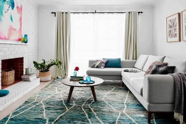 Modern living room with gray sofa, blue-green rug, round wooden table, abstract art, potted plants, and bright window.