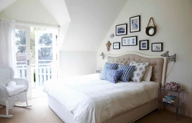 Bright bedroom with a white bed, blue-patterned pillows, wall art, and a wicker chair by glass doors leading to a balcony.