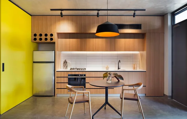 Modern kitchen with wooden cabinets, yellow wall, small dining table, chairs, and pendant light.