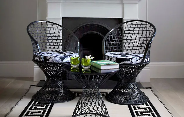 Two black wire chairs with patterned cushions, a round table with books and green glasses in a room with a fireplace.
