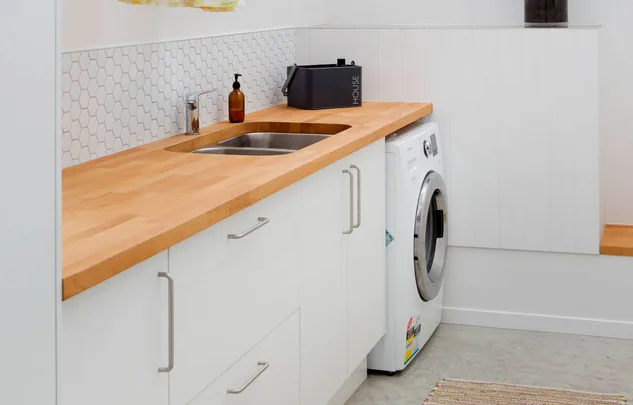 Laundry room with wooden countertop, sink, washing machine, and white cabinets.