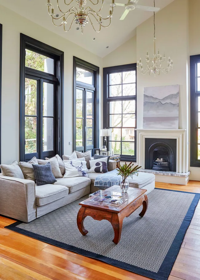 Stylish living room with beige sofa, large windows, chandeliers, and a fireplace, featuring modern decor and wood accents.
