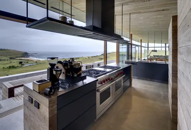 Modern kitchen with sleek appliances, large windows, and stunning coastal view with green landscape and ocean.