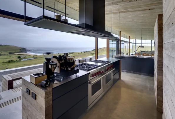 Modern kitchen with sleek appliances, large windows, and stunning coastal view with green landscape and ocean.