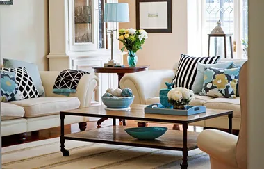 Bright living room with white sofas, blue and floral cushions, wooden table, and decorative items. Natural light fills the room.