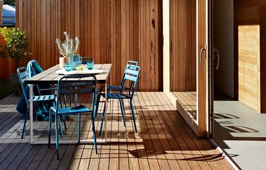 Outdoor patio with wooden deck, blue metal chairs, a table with glassware, and potted plants, against a wooden wall.
