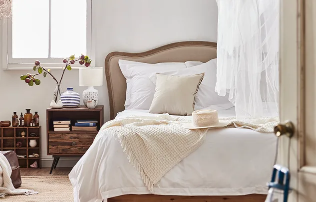 Cozy bedroom with white bedding, a beige headboard, wooden nightstand, and decorative items on a sunny morning.