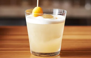 A glass of whiskey sour cocktail on a wooden table, garnished with a cherry and ice cube.