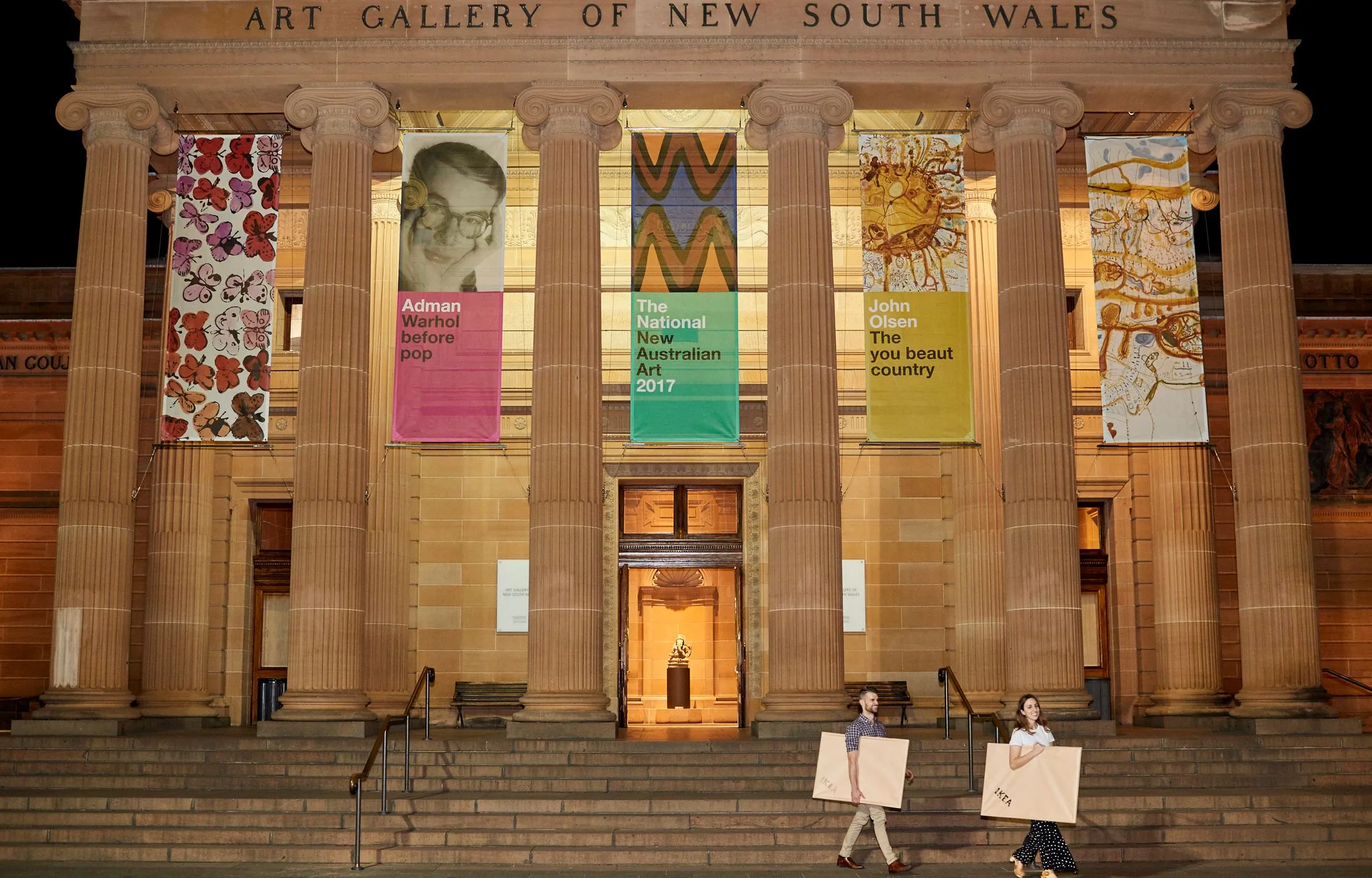 Two people carry art in front of the Art Gallery of New South Wales, with banners for Andy Warhol and John Olsen exhibits.