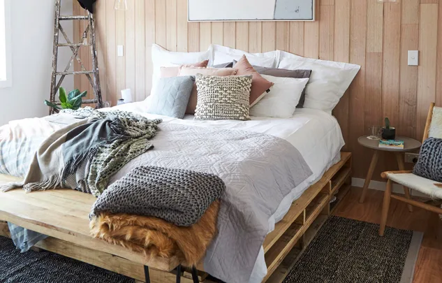 Cozy bedroom with a pallet bed, layered pillows, throws, wooden walls, and rustic decor.