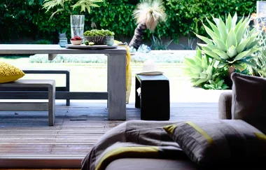 Outdoor dining area with a wooden table, bench, and fresh greenery, set against a lush garden background.