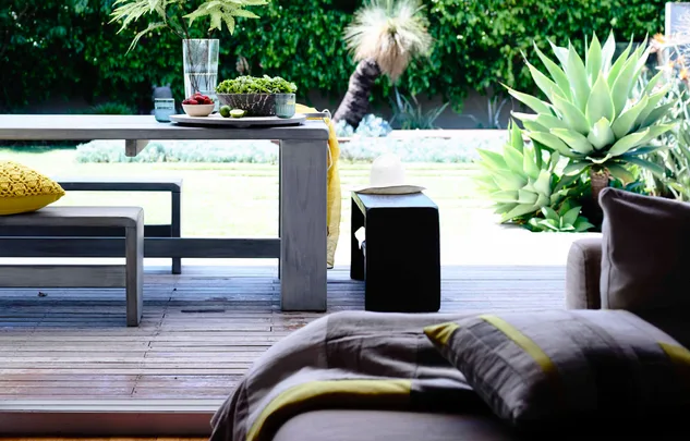 Outdoor dining area with a wooden table, bench, and fresh greenery, set against a lush garden background.