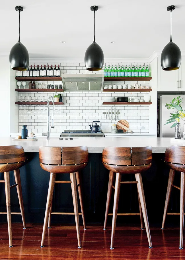 Modern kitchen with wooden bar stools, white subway tiles, open shelves, and black pendant lights.