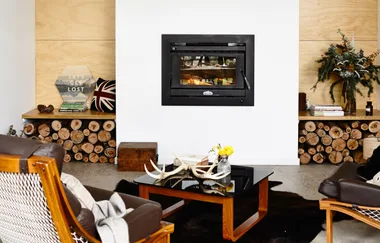 Cozy living room with modern fireplace, leather chairs, and a black coffee table with antlers and flowers. Logs stored nearby.