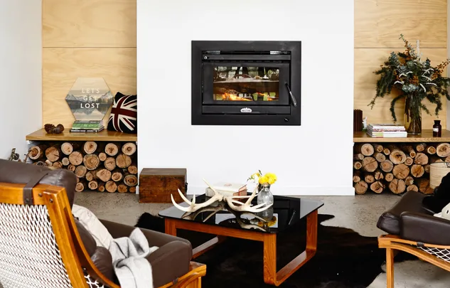 Cozy living room with modern fireplace, leather chairs, and a black coffee table with antlers and flowers. Logs stored nearby.