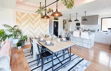 Modern dining and kitchen area with wooden table, black chairs, pendant lights, and potted plants.