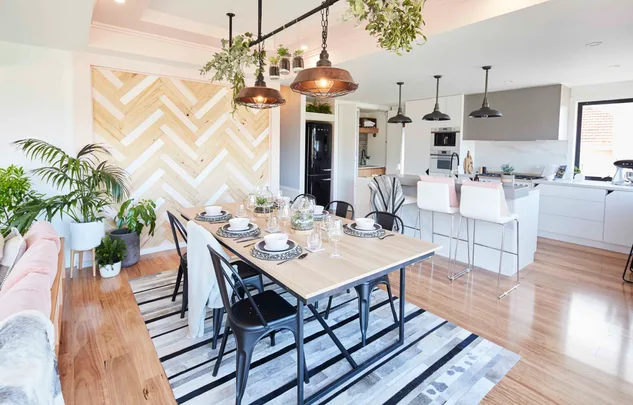 Modern dining and kitchen area with wooden table, black chairs, pendant lights, and potted plants.