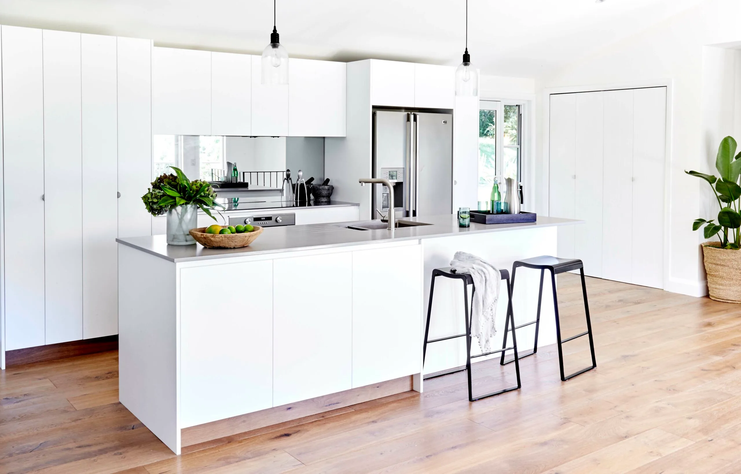 Modern white kitchen with island, two bar stools, pendant lights, and wooden floor. Green plant decor on the counter.