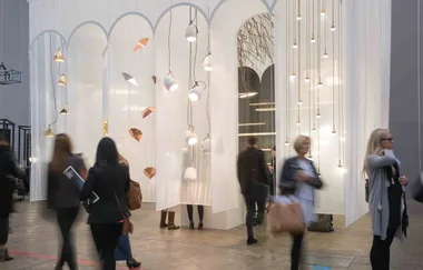 Exhibition space with modern pendant lights and people walking, set against white sheer curtains with geometric designs.