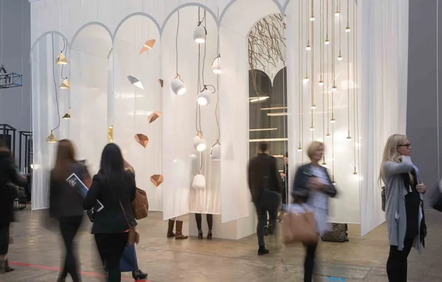 Exhibition space with modern pendant lights and people walking, set against white sheer curtains with geometric designs.