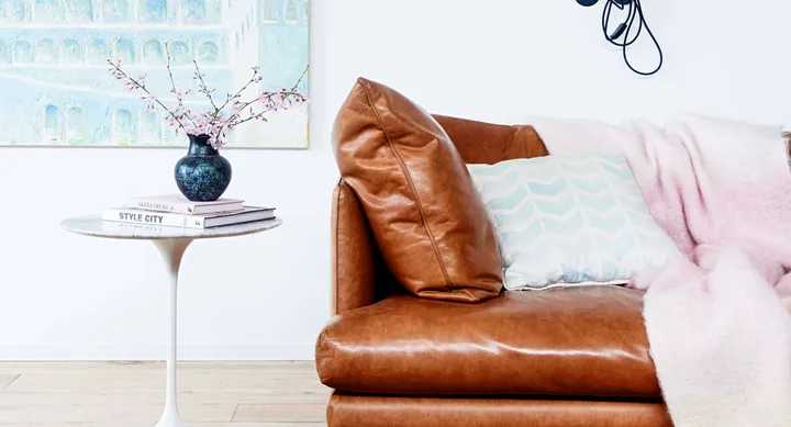 Brown leather couch with light blue pillow and throw, next to a round table with books and a vase of flowers.