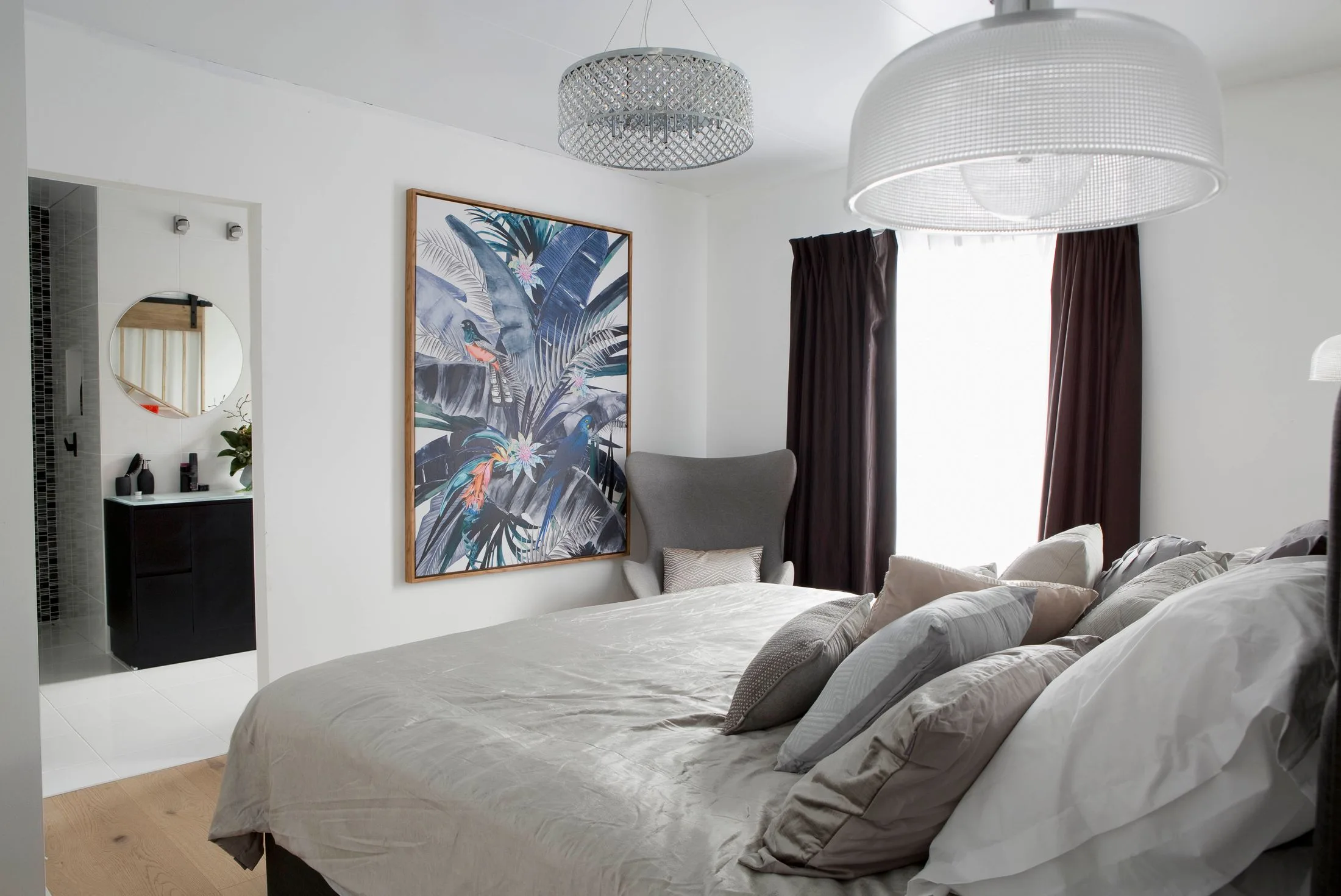 Bedroom with beige bedding, large tropical print artwork, chair, and view into modern bathroom with round mirror.