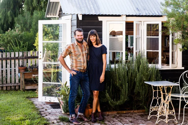 A couple stands smiling in front of a rustic garden shed, surrounded by greenery and a vintage patio table.