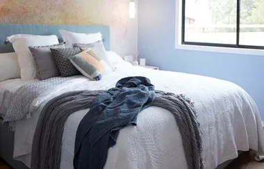 Cozy bedroom with a neatly made bed, layered gray and blue blankets, assorted patterned pillows, and a large window.