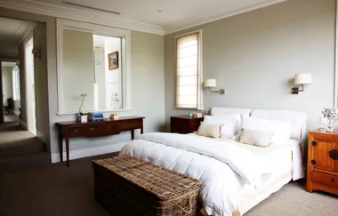 Modern bedroom with white bedding, wicker trunk, wooden furniture, large window, and wall sconces.